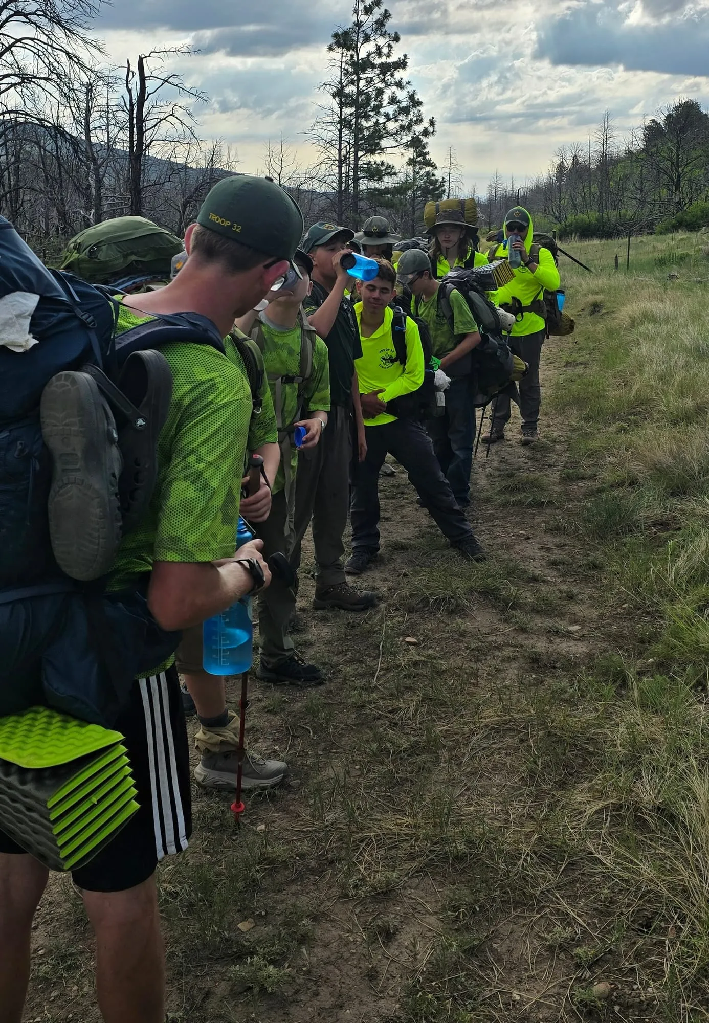 A line of Scouts carrying backpacks pauses on a mountain trail to drink water under a cloudy sky.