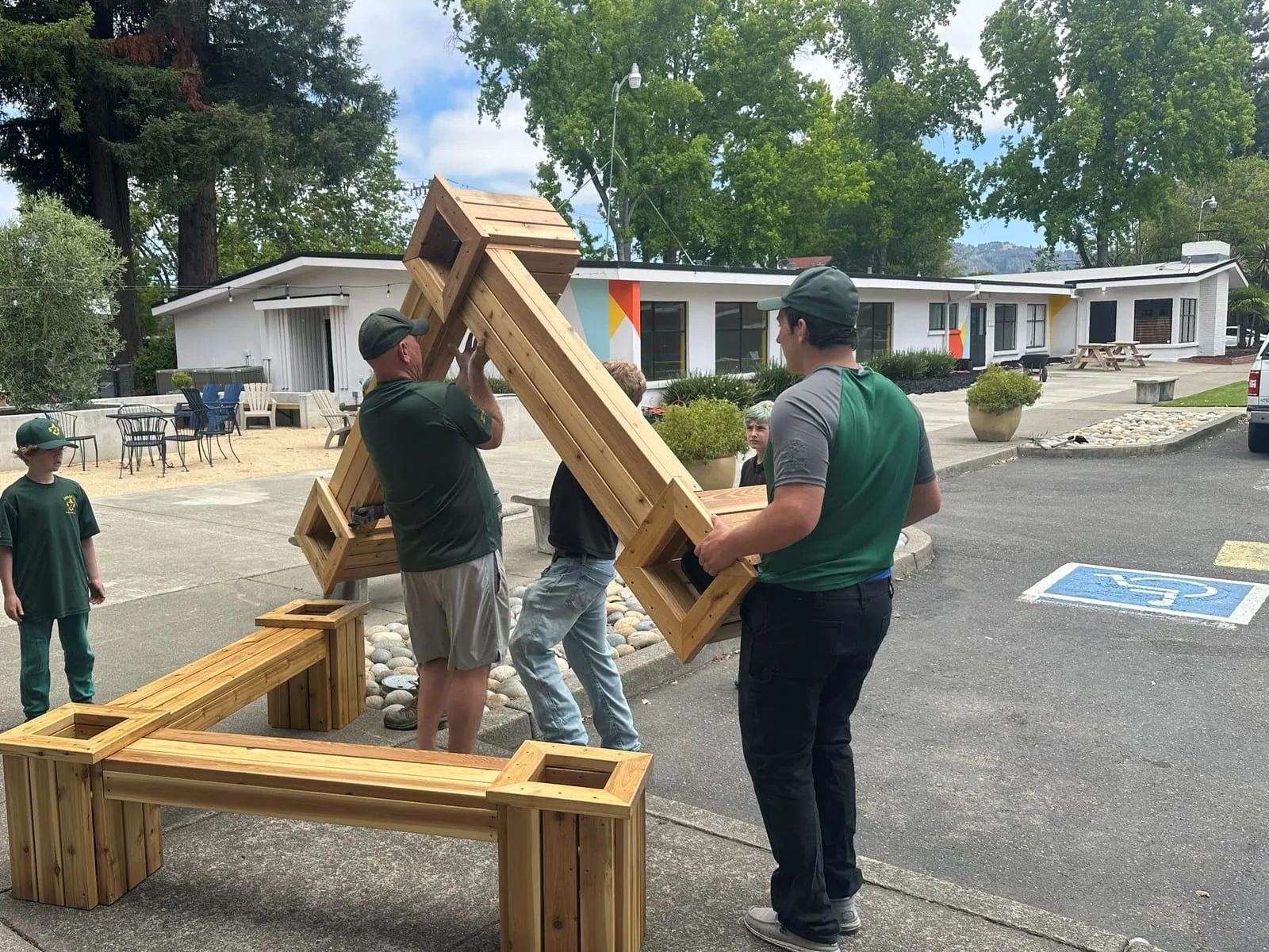 Scouts and an adult volunteer lift a newly built wooden planter bench into place in a courtyard.