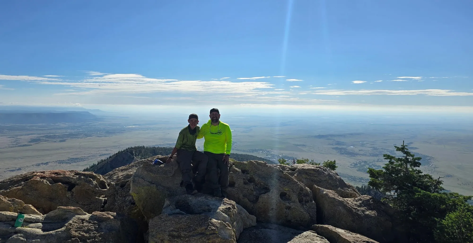A Scout and an adult leader sit on a rocky mountain summit above a wide plain that stretches to the horizon.