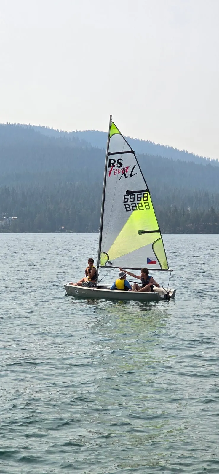 Scouts in life jackets sailing a small sailboat across a wide, forested lake.